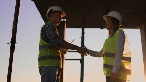 Builders-man-with-a-tablet-and-a-woman-in-white-helmets-shake-hands-at-sunset-standing-on-the-roof-of-the-building